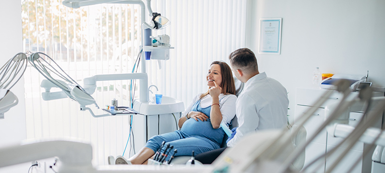 Femme enceinte assise dans un fauteuil dentaire et discutant avec un praticien - La Prévention Médicale Femme enceinte assise dans un fauteuil dentaire et discutant avec un praticien - La Prévention Médicale
