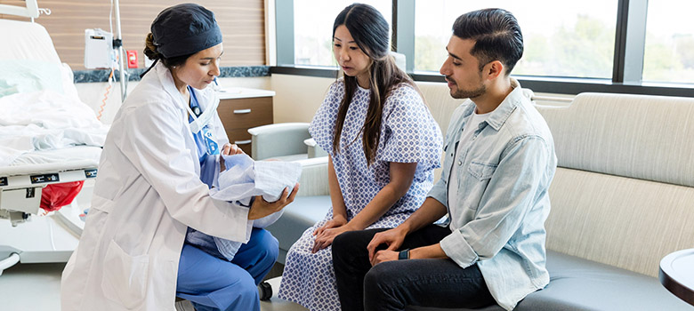 Une professionnelle de santé tenant un nouveau-né dans les bras discute avec les parents dans la chambre de l'hôpital - La Prévention Médicale Une professionnelle de santé tenant un nouveau-né dans les bras discute avec les parents dans la chambre de l'hôpital - La Prévention Médicale