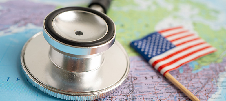 Un stéthoscope posé sur une carte du monde avec le drapeau américain miniature - La Prévention Médicale Un stéthoscope posé sur une carte du monde avec le drapeau américain miniature - La Prévention Médicale