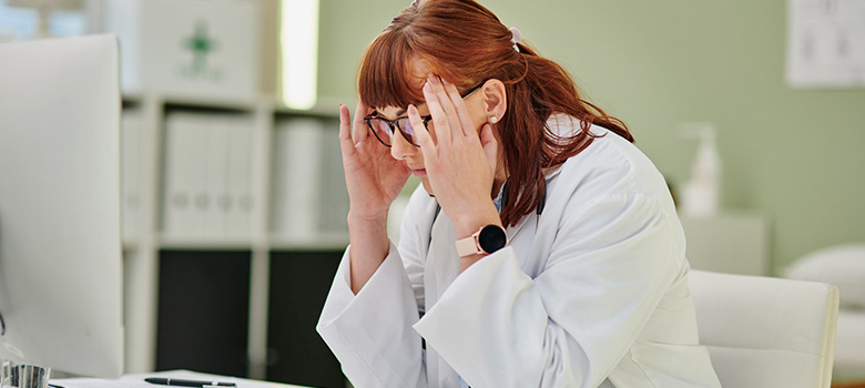 Une femme médecin, assise à son bureau, se tient la tête devant l'écran de son ordinateur - La Prévention Médicale